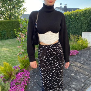 K&ouml;nnte beinhalten: Ein schwarzer Rollkragenpullover mit einem wei&szlig;en Korsett und einem schwarz-wei&szlig;en Polka-Dot-Rock. Die Person tr&auml;gt eine schwarze Sonnenbrille.