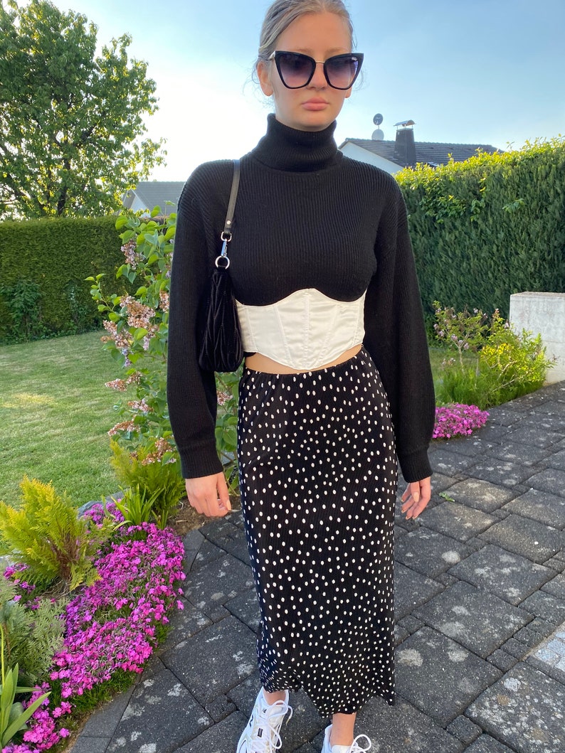 K&ouml;nnte beinhalten: Ein schwarzer Rollkragenpullover mit einem wei&szlig;en Korsett und einem schwarz-wei&szlig;en Polka-Dot-Rock. Die Person tr&auml;gt eine schwarze Sonnenbrille.