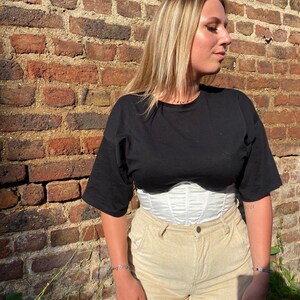 K&ouml;nnte beinhalten: Eine Frau tr&auml;gt ein schwarzes T-Shirt mit einem wei&szlig;en Korsett und einer beigefarbenen Cordhose. Sie steht vor einer Backsteinmauer.
