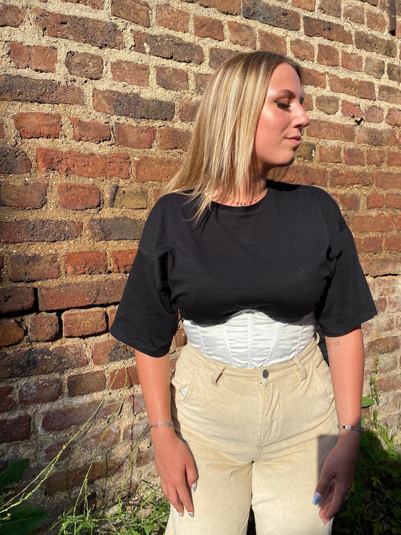 K&ouml;nnte beinhalten: Eine Frau tr&auml;gt ein schwarzes T-Shirt mit einem wei&szlig;en Korsett und einer beigefarbenen Cordhose. Sie steht vor einer Backsteinmauer.