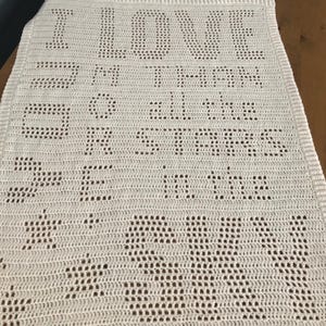 May include: A white crocheted blanket with the words "I LOVE THEM MORE THAN ALL THE STARS IN THE SKY" stitched into the fabric. The blanket is rectangular and has a textured surface.