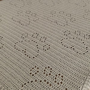 Pode incluir: Um close-up de um cobertor de crochê de cor cinza claro com um padrão de losango repetido.