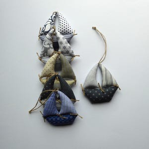 Könnte beinhalten: Eine Sammlung handgefertigter Segelboot-Ornamente aus Stoff in verschiedenen Mustern und Farben, darunter Blau, Weiß und Grau. Jedes Boot hat eine kleine Schlaufe zum Aufhängen und ist mit Bindfaden gebunden. Die Segelboote sind auf einer weißen Oberfläche angeordnet.