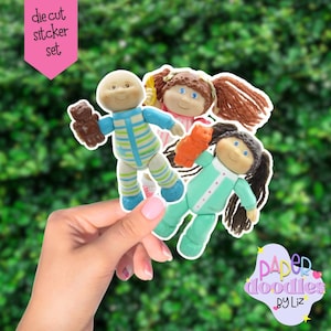 May include: A die-cut sticker set featuring three Cabbage Patch Kids dolls. One doll is in blue and green striped pajamas, holding a brown bear. Another doll is in pink and white pajamas, and the third doll is in green pajamas.