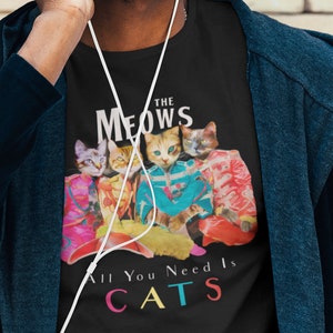 K&ouml;nnte beinhalten: Ein schwarzes T-Shirt mit einem Motiv von vier Katzen in bunten Outfits. Der Text "The Meows" befindet sich &uuml;ber den Katzen und "All You Need Is Cats" unter den Katzen. Der Text ist in einer bunten Schriftart.