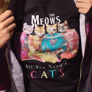 K&ouml;nnte beinhalten: Eine Frau tr&auml;gt eine schwarze Jacke mit Blumendruck und ein schwarzes T-Shirt mit einem Katzenmotiv und dem Text "The Meows All You Need Is Cats".