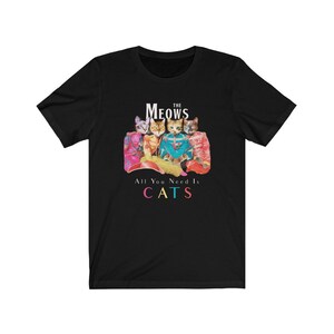 K&ouml;nnte beinhalten: Schwarzes T-Shirt mit einem Motiv von vier Katzen in bunten Outfits. Der Text "The Meows" befindet sich &uuml;ber den Katzen und "All You Need Is Cats" darunter.