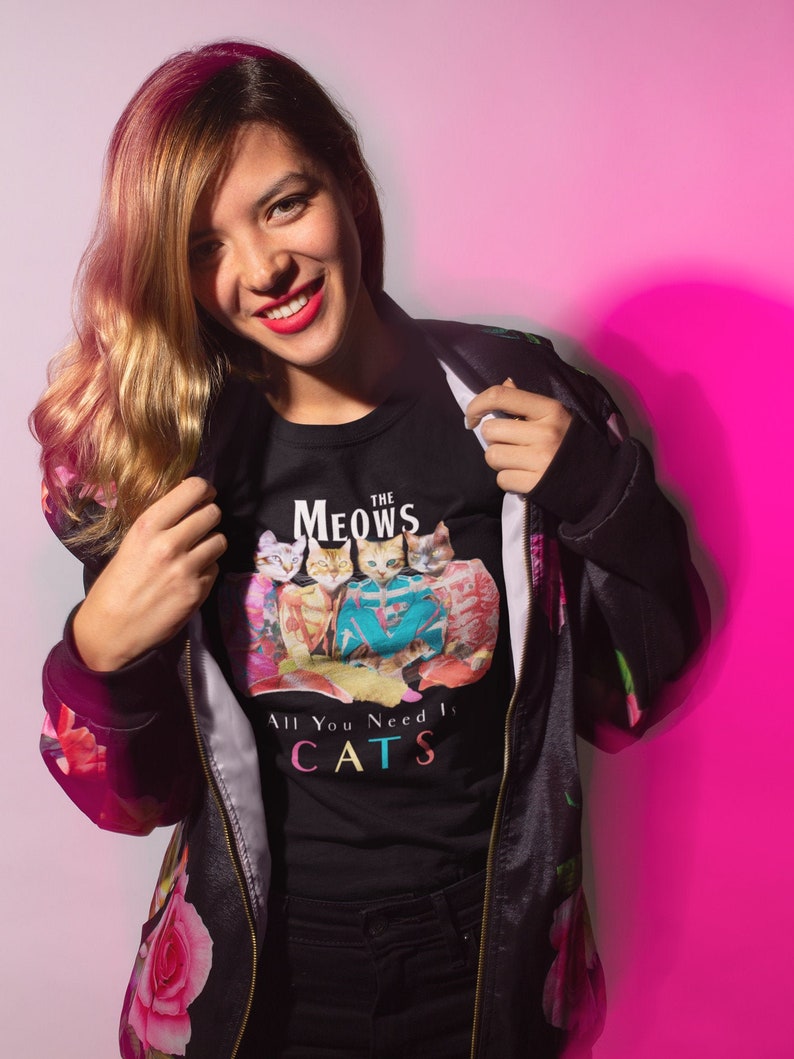 K&ouml;nnte beinhalten: Eine Frau tr&auml;gt eine schwarze Jacke mit Blumendruck und ein schwarzes T-Shirt mit einem Katzenmotiv und dem Text "The Meows All You Need Is Cats".