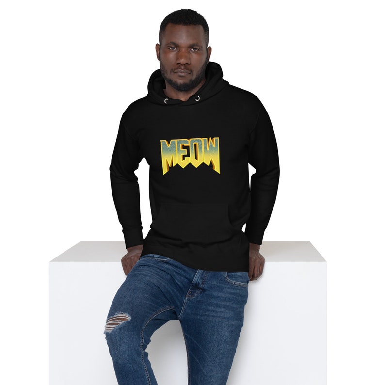 Pu&ograve; includere: Una felpa con cappuccio nera con una grafica "MEOW" gialla e blu sul davanti. La felpa con cappuccio &egrave; indossata da una persona con la barba e si trova su una superficie bianca.