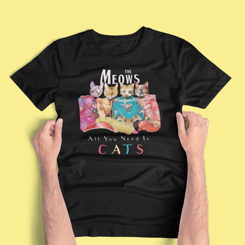 K&ouml;nnte beinhalten: Schwarzes T-Shirt mit einem Motiv von vier Katzen in bunten Outfits. Der Text "The Meows" befindet sich &uuml;ber den Katzen und "All You Need Is CATS" darunter.