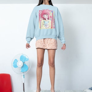 May include: A light blue sweatshirt with a graphic print of an anime character holding a cat. The text on the sweatshirt says "きみとあいたい".
