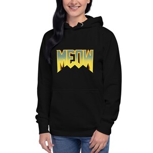 Pu&ograve; includere: Felpa con cappuccio nera con una grafica "MEOW" gialla e arancione in uno stile di font da videogioco retr&ograve;.