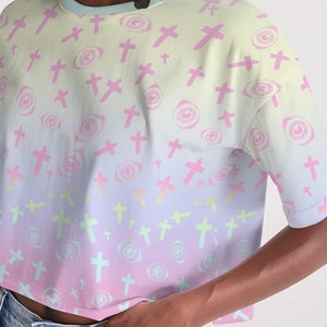 May include: A pastel pink, blue and yellow cropped t-shirt with a repeating pattern of pink crosses and stylised eyes. The shirt has a relaxed fit and short sleeves.