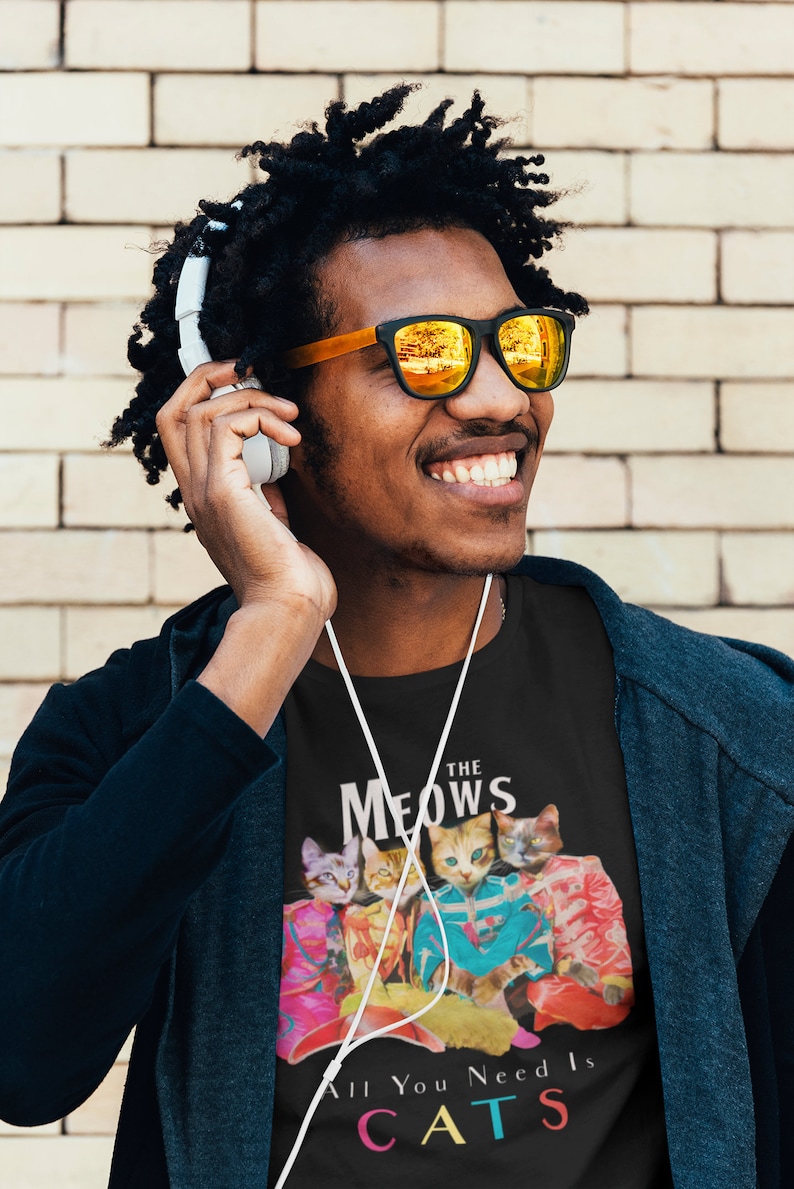 K&ouml;nnte beinhalten: Ein schwarzes T-Shirt mit einem Motiv von vier Katzen in bunten Outfits. Der Text "The Meows" befindet sich &uuml;ber den Katzen und "All You Need Is Cats" unter den Katzen. Der Text ist in einer bunten Schriftart.