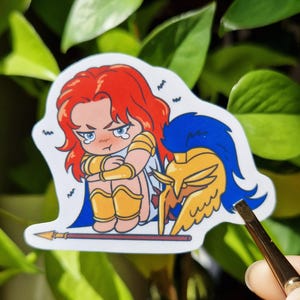 Può includere: Un adesivo con un'illustrazione a cartoni animati di una guerriera abbattuta con capelli rossi, armatura dorata e mantello blu. Un elmo d'oro e una lancia sono anch'essi presenti. La guerriera è seduta con le braccia attorno alle ginocchia, con un'espressione triste.