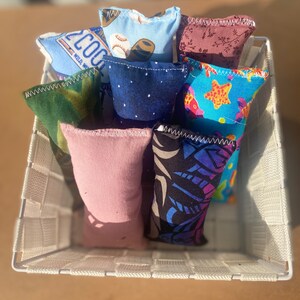 May include: A white wicker basket filled with eight colorful fabric sensory bags. The bags have different patterns, including a baseball, a starfish, and a camouflage design.