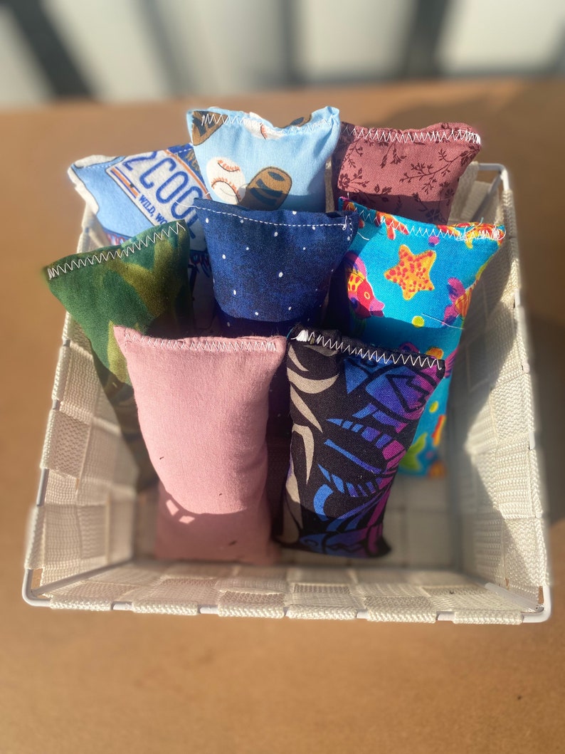 May include: A white wicker basket filled with eight colorful fabric sensory bags. The bags have different patterns, including a baseball, a starfish, and a camouflage design.