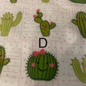 May include: A light pink fabric featuring a repeating pattern of cartoon cacti. The cacti are various shades of green with smiling faces and small pink flowers. Some cacti have a pink bow. The letter "D" is printed in black.