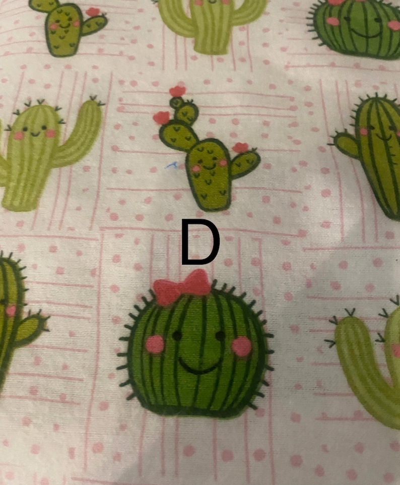 May include: A light pink fabric featuring a repeating pattern of cartoon cacti. The cacti are various shades of green with smiling faces and small pink flowers. Some cacti have a pink bow. The letter "D" is printed in black.