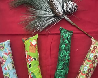 May include: Four festive catnip toys with different holiday patterns. The toys are green, red, blue, and brown with a variety of holiday designs. The toys are on a red surface with a green pine branch with snow.