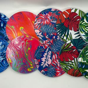 May include: Eight round coasters with tropical leaf designs. The coasters feature a variety of colors, including red, blue, green, and white.