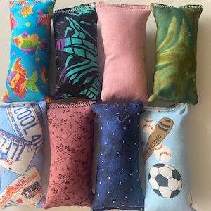 Puede incluir: Nueve bolsitas de tela coloridas con diferentes patrones. Las bolsitas están llenas de frijoles o arroz y están diseñadas para ser usadas como pelotas antiestrés o juguetes sensoriales. Los patrones incluyen una bolsita azul con peces, una bolsita negra y verde con un patrón de cebra, una bolsita rosa, una bolsita verde con un patrón de hoja, una bolsita azul con una placa de matrícula, una bolsita rosa con un patrón floral, una bolsita azul con un patrón estrellado, una bolsita azul con un bate de béisbol y una pelota de béisbol, y una bolsita azul con una pelota de fútbol.