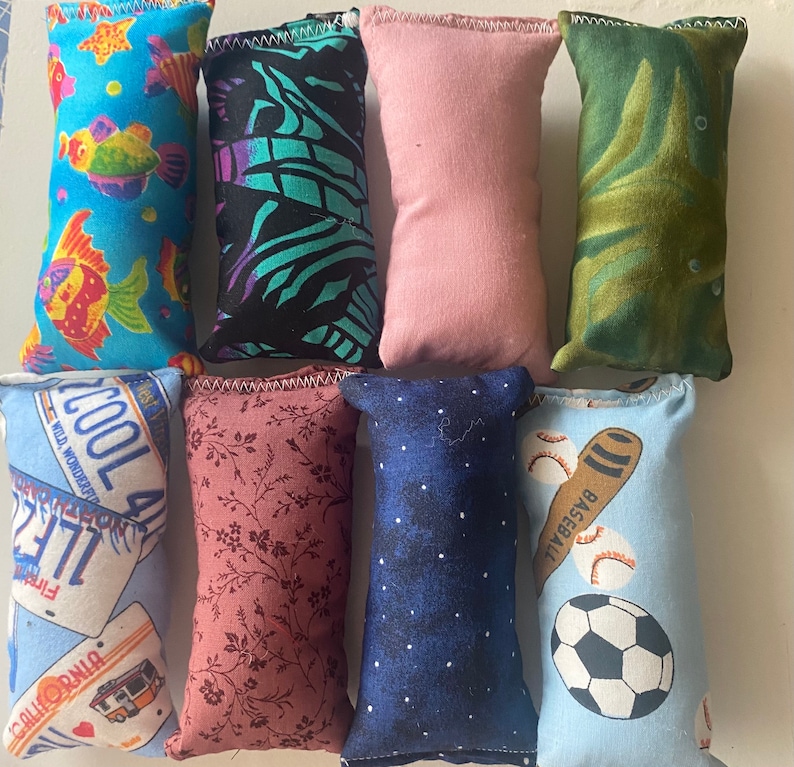 May include: Nine colorful fabric pouches with different patterns. The pouches are filled with beans or rice and are designed to be used as stress balls or sensory toys. The patterns include a blue pouch with fish, a black and green pouch with a zebra pattern, a pink pouch, a green pouch with a leaf pattern, a blue pouch with a license plate, a pink pouch with a floral pattern, a blue pouch with a starry pattern, a blue pouch with a baseball and bat, and a blue pouch with a soccer ball.