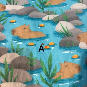 May include: A fabric pattern showing capybaras in a blue river with grey rocks, green plants, and orange lily pads. The letter "A" is printed in black on the fabric. The capybaras are light brown.