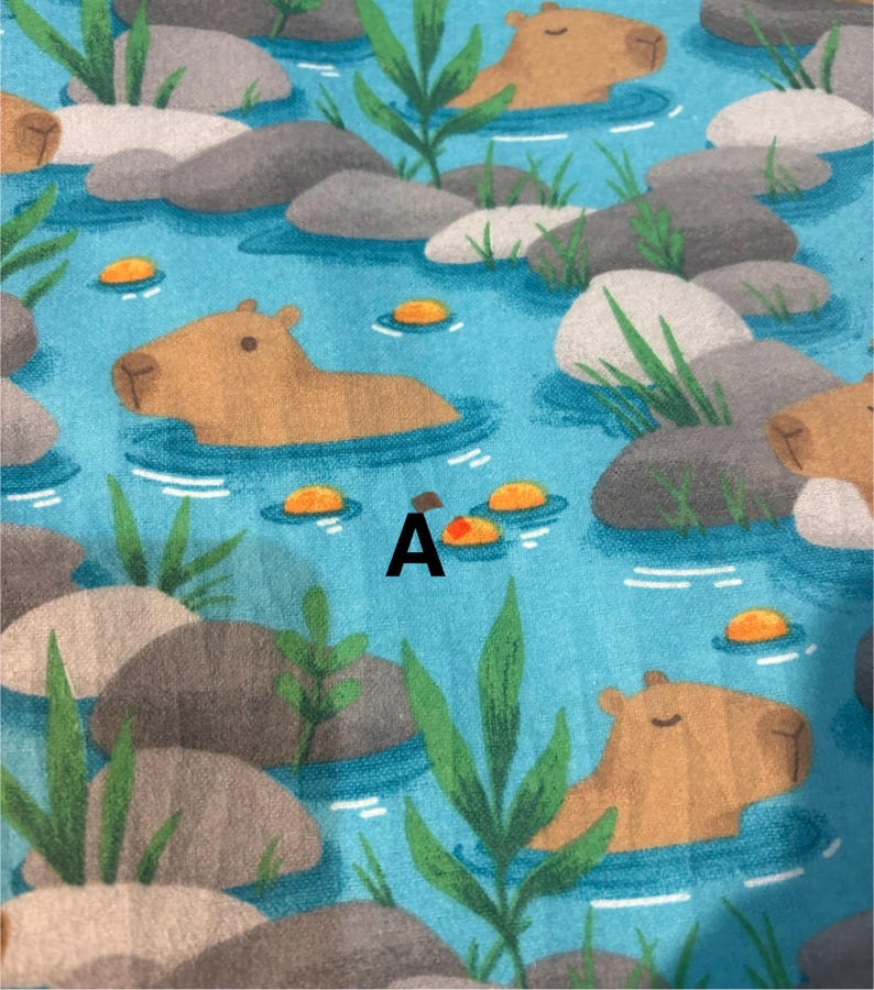 May include: A fabric pattern showing capybaras in a blue river with grey rocks, green plants, and orange lily pads. The letter "A" is printed in black on the fabric. The capybaras are light brown.