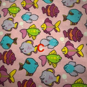 May include: A pink fabric featuring a repeating pattern of cartoon fish. The fish are in various colours, including blue, purple, green, and orange, with yellow fins and pink tails. The fish have smiling faces.