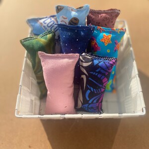 May include: A white wicker basket filled with eight colorful fabric catnip toys. The toys are made of different fabrics with various patterns, including stars, fish, and geometric designs.