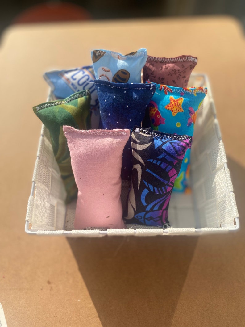 May include: A white wicker basket filled with eight colorful fabric catnip toys. The toys are made of different fabrics with various patterns, including stars, fish, and geometric designs.