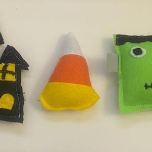 May include: Three felt Halloween-themed toys: a black haunted house, a yellow and orange candy corn, and a green Frankenstein monster.
