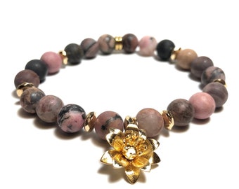 Puede incluir: Un brazalete de piedras preciosas rosa y gris con detalles dorados y un dije de flor dorada.