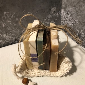 May include: A set of six handmade soap bars wrapped in twine and placed in a small white mesh bag. The soap bars are in various colors, including brown, beige, and green.