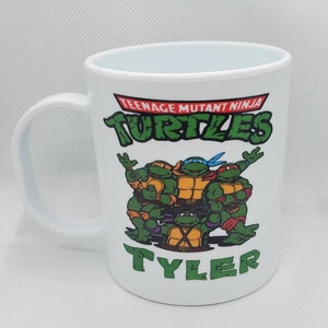 May include: White ceramic mug featuring the Teenage Mutant Ninja Turtles. The mug has a green and orange graphic of the turtles, with the text "TEENAGE MUTANT NINJA TURTLES" and the name "TYLER" in green.