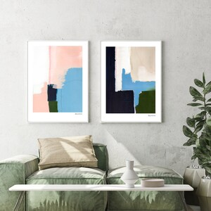 Op de afbeelding: Twee abstracte schilderijen in witte lijsten, een met roze, blauwe en groene kleuren, de andere met zwarte, blauwe en groene kleuren. De schilderijen hangen aan een witte muur boven een groene bank en een witte salontafel.