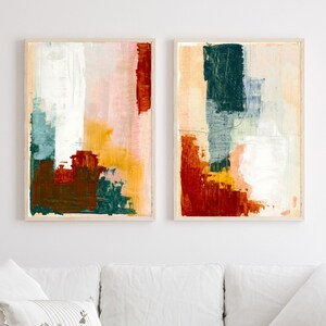 Op de afbeelding: Twee abstracte schilderijen in houten frames. De schilderijen zijn in tinten rood, oranje, geel, groen en wit. De schilderijen hangen aan een witte muur boven een witte bank.