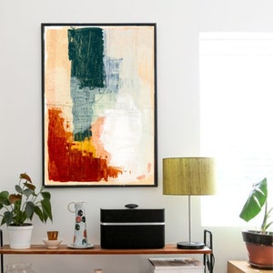 Op de afbeelding: Abstract schilderij met een zwarte lijst, met een mix van rode, oranje, gele, groene en witte verfstreken. Het schilderij hangt aan een witte muur boven een houten plank met een lamp, een speaker en een vaas.
