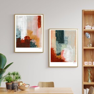 Op de afbeelding: Twee abstracte schilderijen met een witte achtergrond en kleurrijke geometrische vormen in tinten rood, oranje, groen en blauw. De schilderijen zijn ingelijst in goud en hangen aan een witte muur boven een houten eettafel.