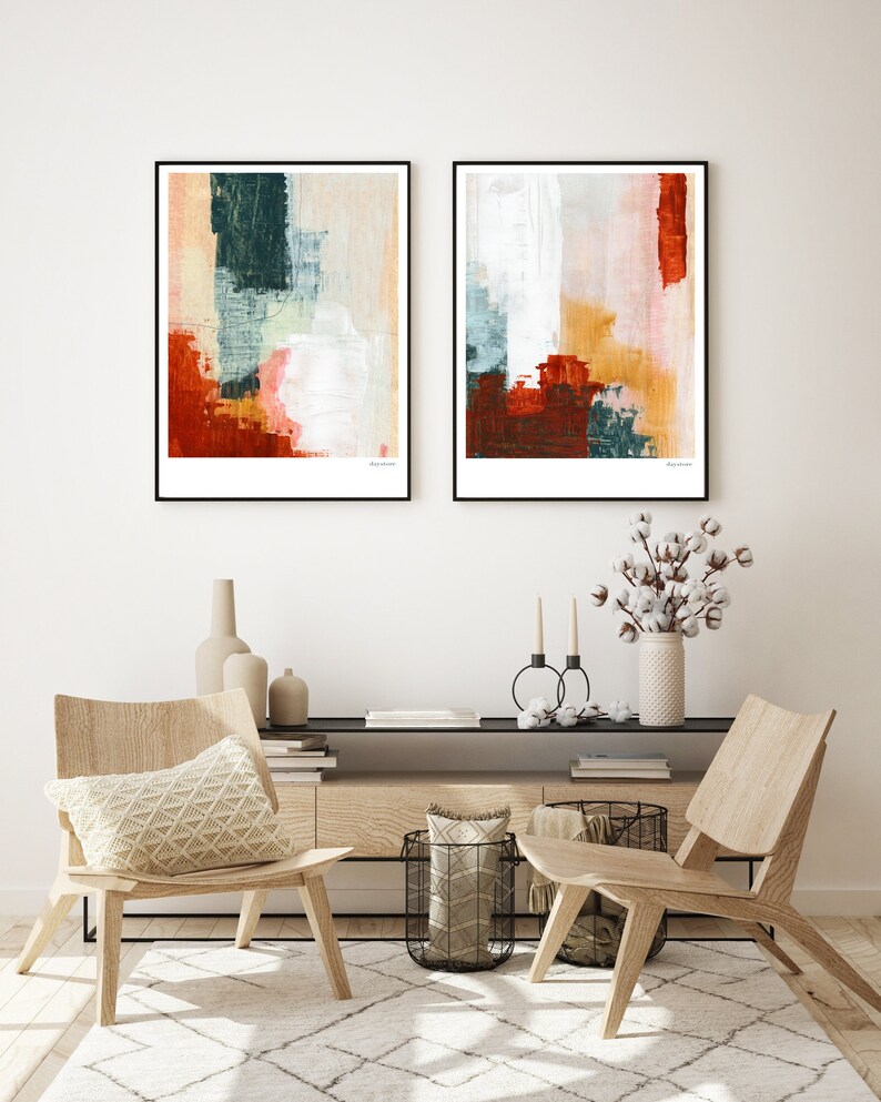 Op de afbeelding: Twee abstracte schilderijen in zwarte lijsten met een witte achtergrond en kleurrijke geometrische vormen in tinten rood, oranje, blauw en groen. De schilderijen hangen aan een witte muur boven een houten consoletafel met twee stoelen.