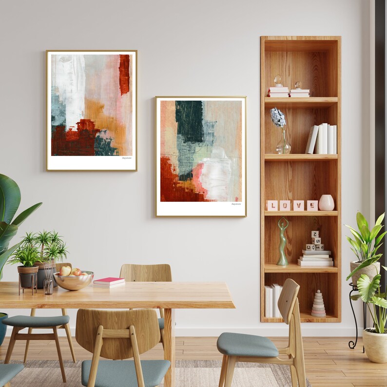 Op de afbeelding: Twee abstracte schilderijen met een witte achtergrond en kleurrijke geometrische vormen in tinten rood, oranje, groen en blauw. De schilderijen zijn ingelijst in goud en hangen aan een witte muur boven een houten eettafel.