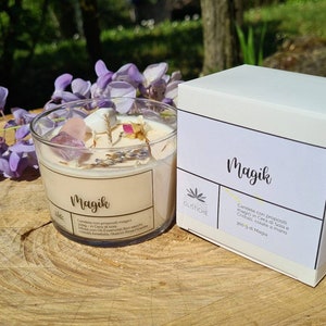 May include: A white candle jar with the word "Magik" printed on the label. The candle jar is filled with a white candle and has crystals and dried flowers inside. The candle jar is sitting on a wooden surface next to a white box with the word "Magik" printed on the label.