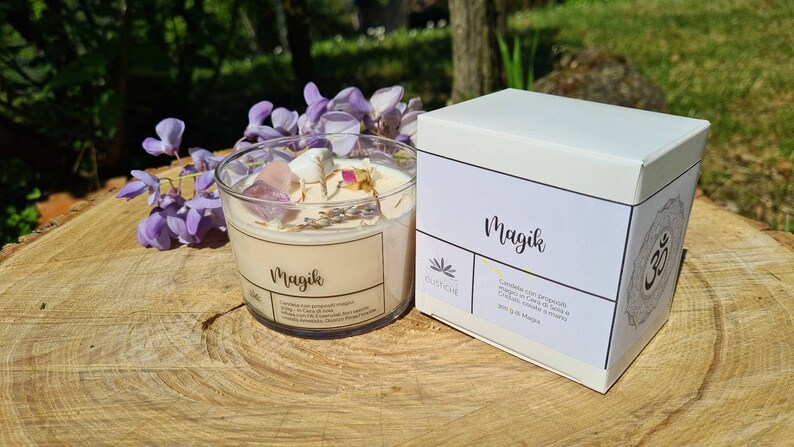 May include: A white candle jar with the word "Magik" printed on the label. The candle jar is filled with a white candle and has crystals and dried flowers inside. The candle jar is sitting on a wooden surface next to a white box with the word "Magik" printed on the label.