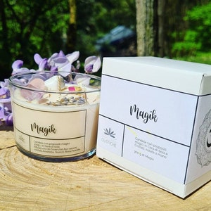 May include: A white candle jar with the word "Magik" printed on the front. The candle is filled with a beige wax and has crystals and dried flowers embedded in the wax. The jar is sitting on a wooden surface. The candle is labeled "Candela con propositi magici 300g in Cera di Soia Infusa con Oli Essenziali, fiori secchi, cristalli Ametista, Quarzo Rosa, Howlite" and "Profezie Olistiche".