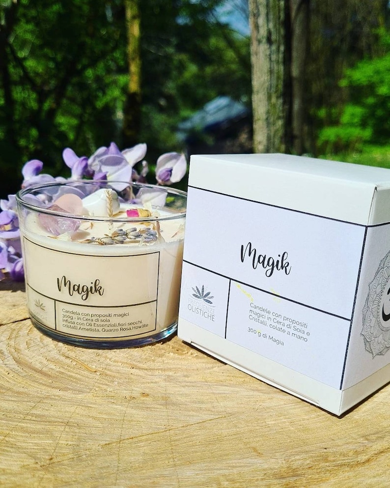 May include: A white candle jar with the word "Magik" printed on the front. The candle is filled with a beige wax and has crystals and dried flowers embedded in the wax. The jar is sitting on a wooden surface. The candle is labeled "Candela con propositi magici 300g in Cera di Soia Infusa con Oli Essenziali, fiori secchi, cristalli Ametista, Quarzo Rosa, Howlite" and "Profezie Olistiche".
