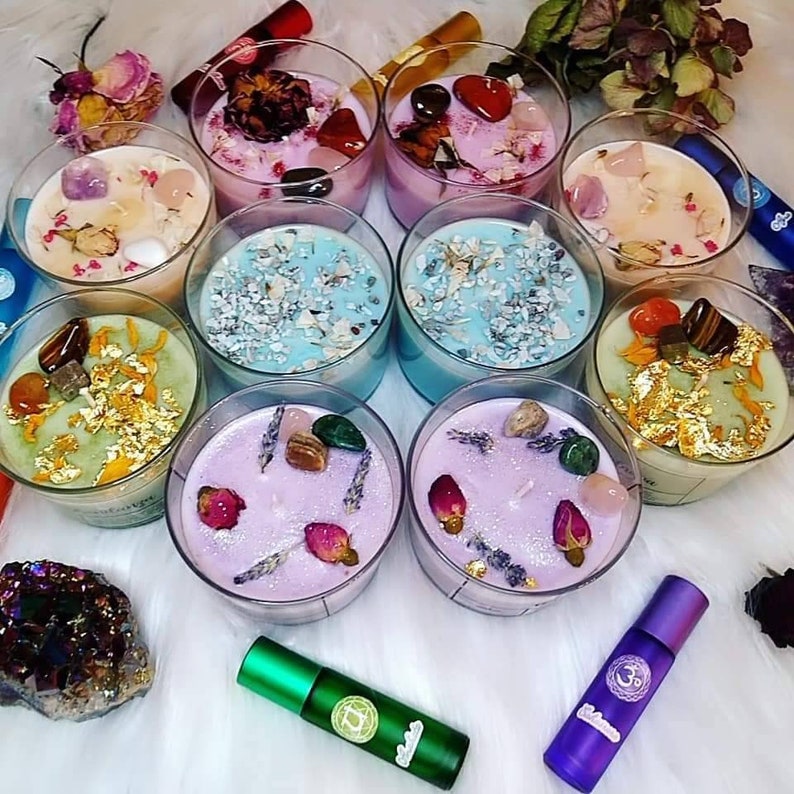 May include: Assortment of colorful candles in glass jars with various crystals, herbs, and other natural elements. The candles are arranged on a white surface with small bottles of essential oils.
