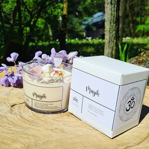 May include: A white candle jar with the word "Magik" printed on the label. The candle is surrounded by crystals and dried flowers. The jar is sitting on a wooden surface. The candle is packaged in a white box with the word "Magik" printed on the label. The box also has a symbol of the Om.