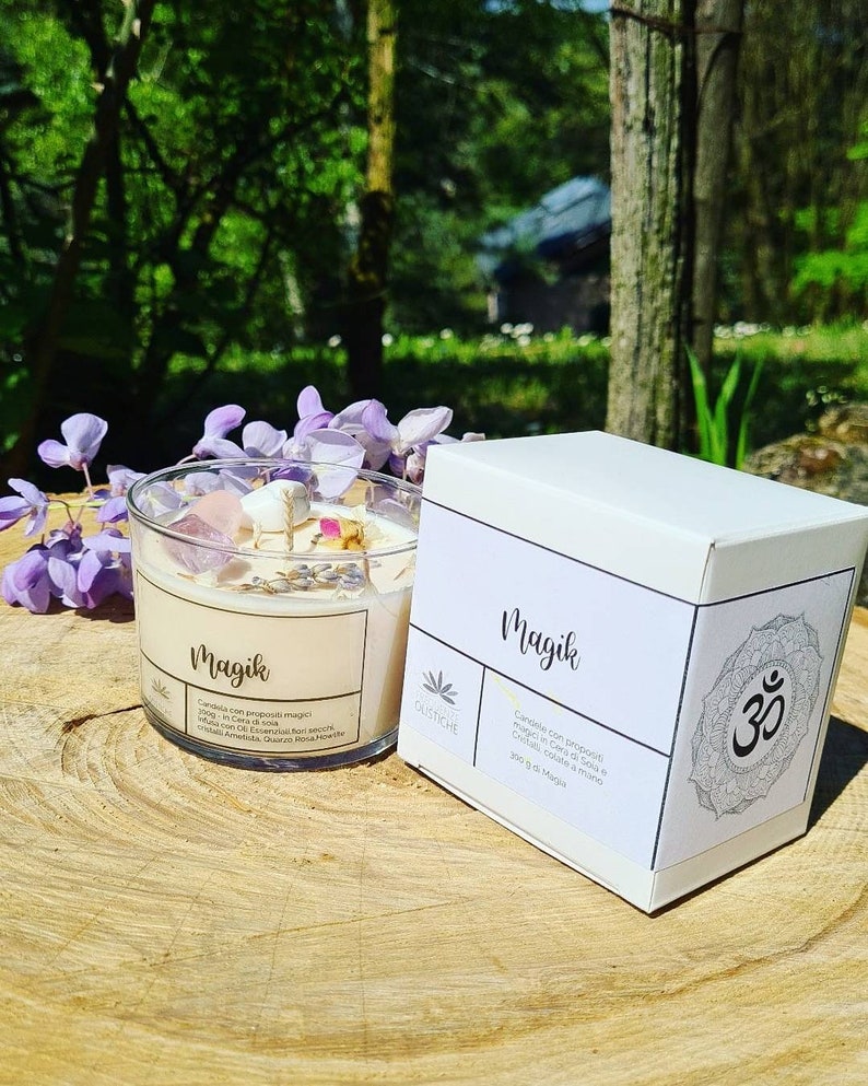 May include: A white candle jar with the word "Magik" printed on the label. The candle is surrounded by crystals and dried flowers. The jar is sitting on a wooden surface. The candle is packaged in a white box with the word "Magik" printed on the label. The box also has a symbol of the Om.