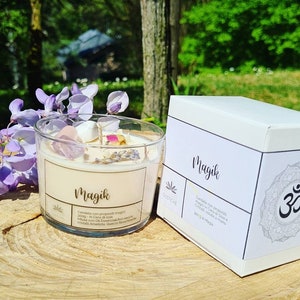 May include: A white candle jar with the word "Magik" printed on the label. The candle is infused with crystals and essential oils. The jar is sitting on a wooden surface with a white box next to it. The box also has the word "Magik" printed on it.